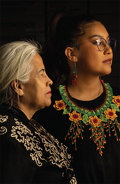 Two women, wearing beadwork embroidered tops photographed facing off to the side