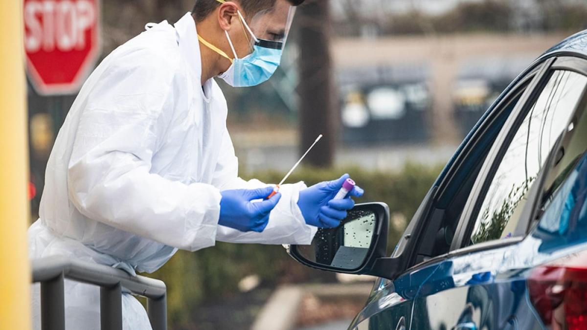 A health care worker in a white coat and PPE puts a bandaid on the arm of a person in a car.