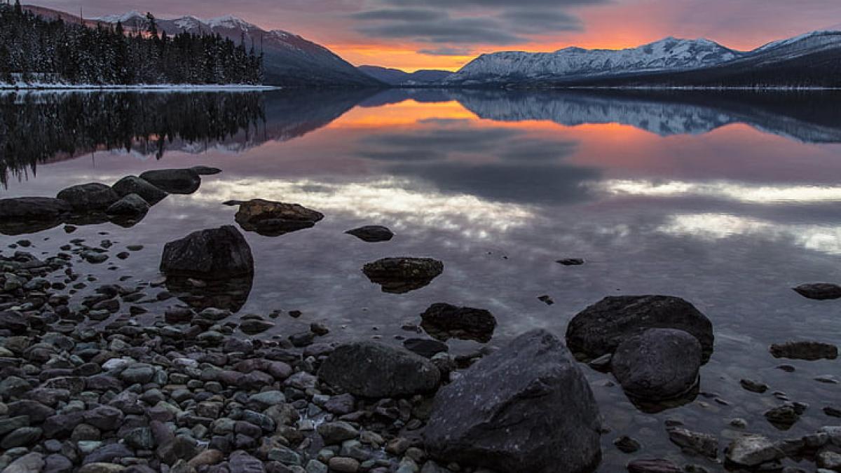 A sunset is reflected in still water with rocks in the foreground and snowy mountains in the distance. 